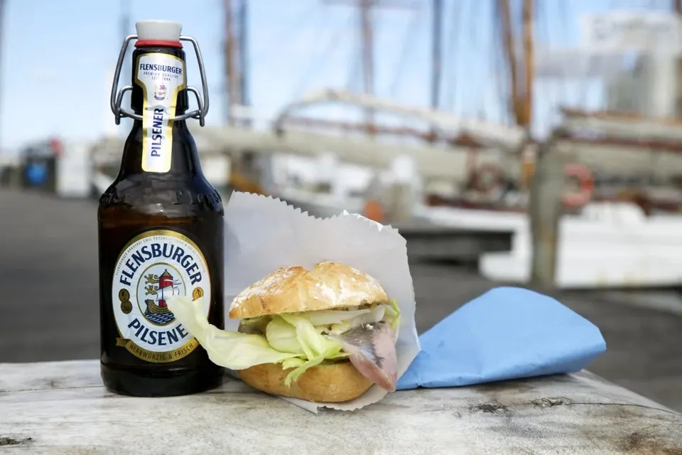 Flensburger Pilsener Bügelflasche und Fischbrötchen am Hafen mit Segelschiff im Hintergrund