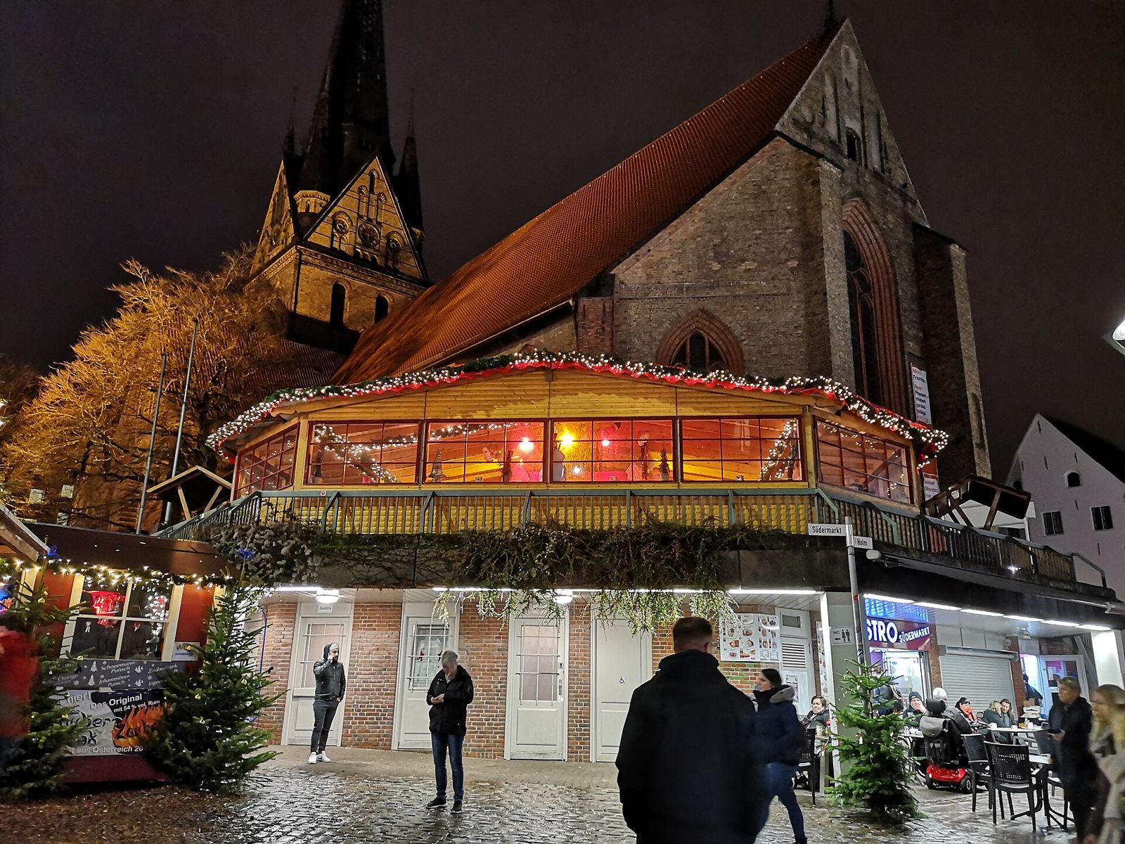 Weihnachtsmarkt in Flensburg am Abend mit beleuchtetem Pavillon, Lichterketten und Tannenbäumen vor der Backsteinkirche